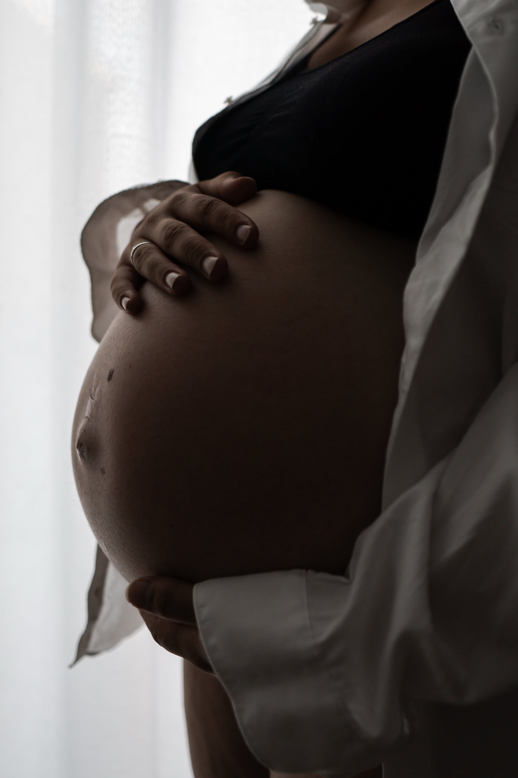 Zwangere vrouw houdt haar buik vast, in een wit overhemd bij zacht raamlicht